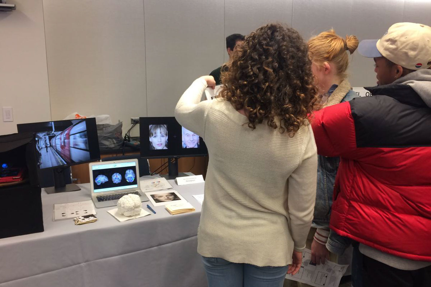 People standing, with their backs turned, at table viewing various printed materials, screens, and a brain model