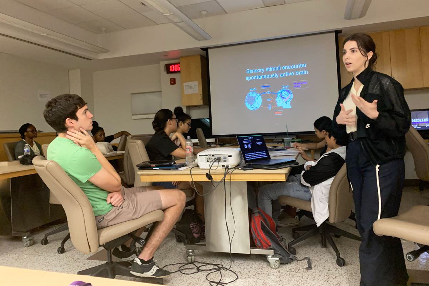 Ella standing and gesturing with her hands, sharing slides on a screen in a dimmed room full students seated at tables