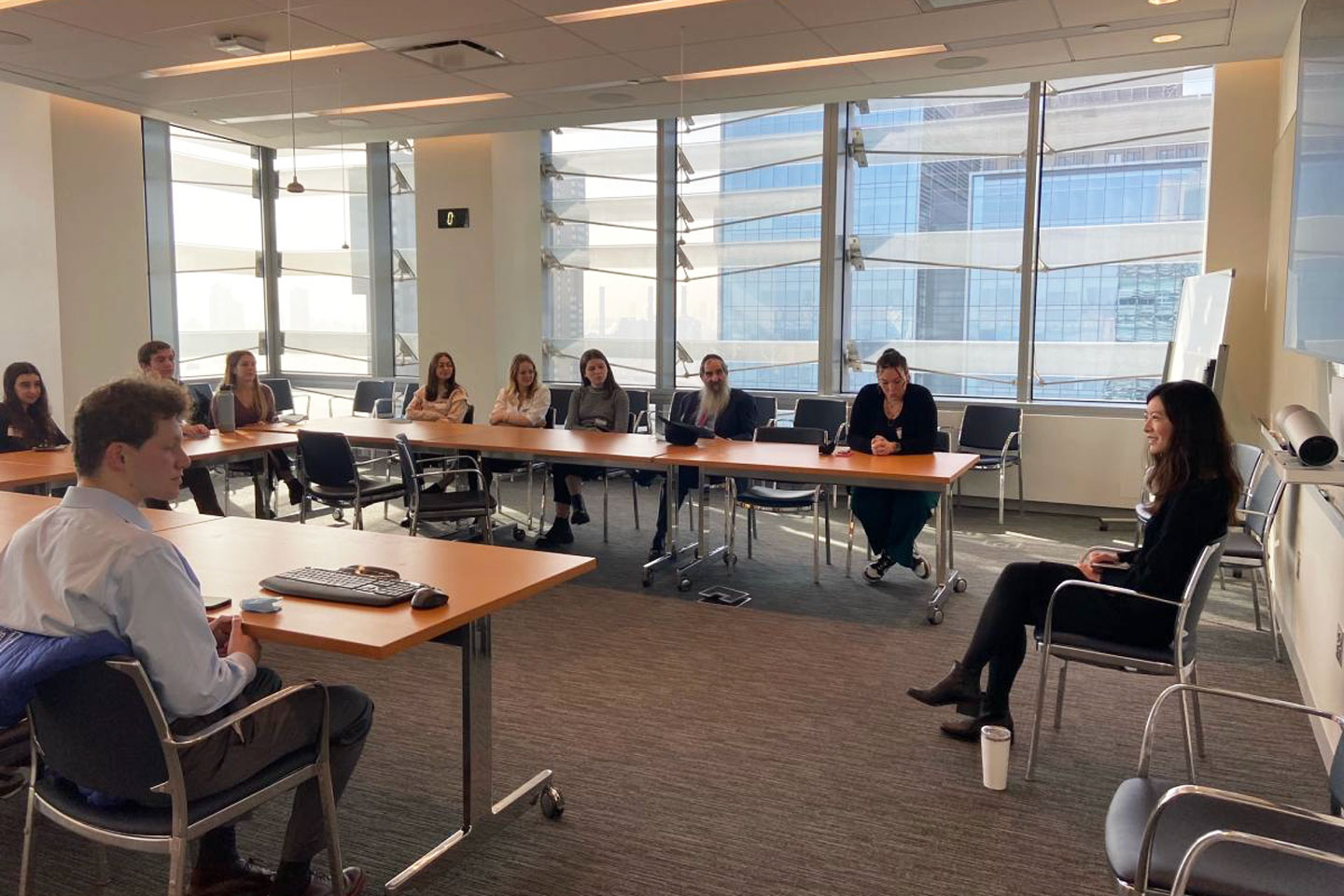 Dr. He smiling and sitting on chair in front of room of people seated around a U-shaped group of tables